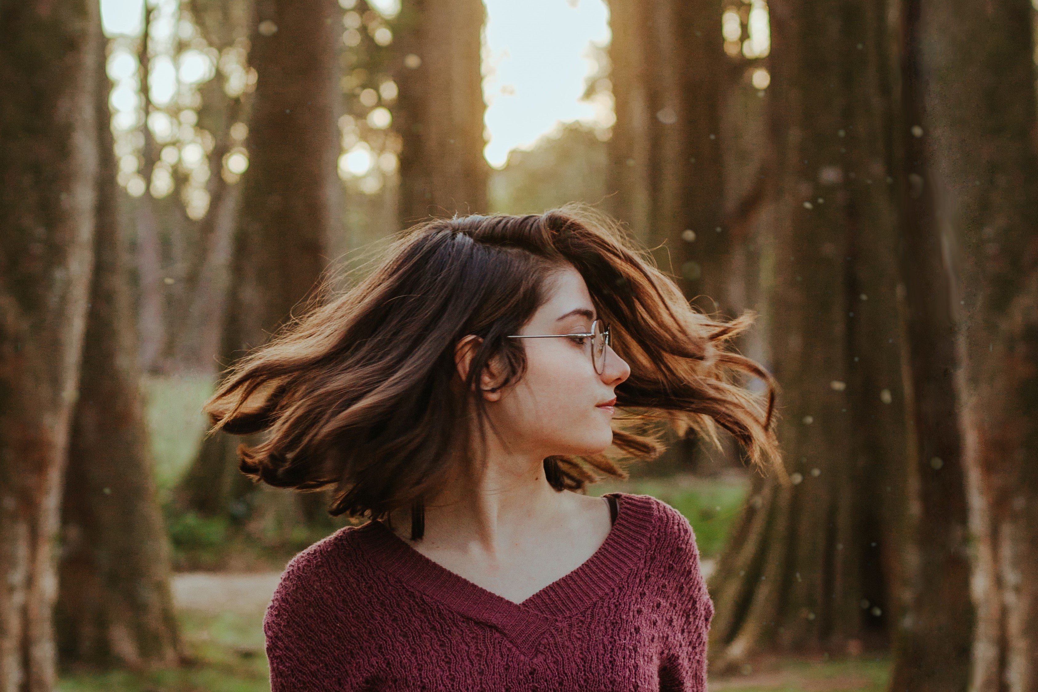 Mulher branca de óculos balançando o cabelo ao vento em uma floresta